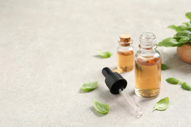 Photo of Bottles of essential basil oil and fresh leaves on light grey table, space for text