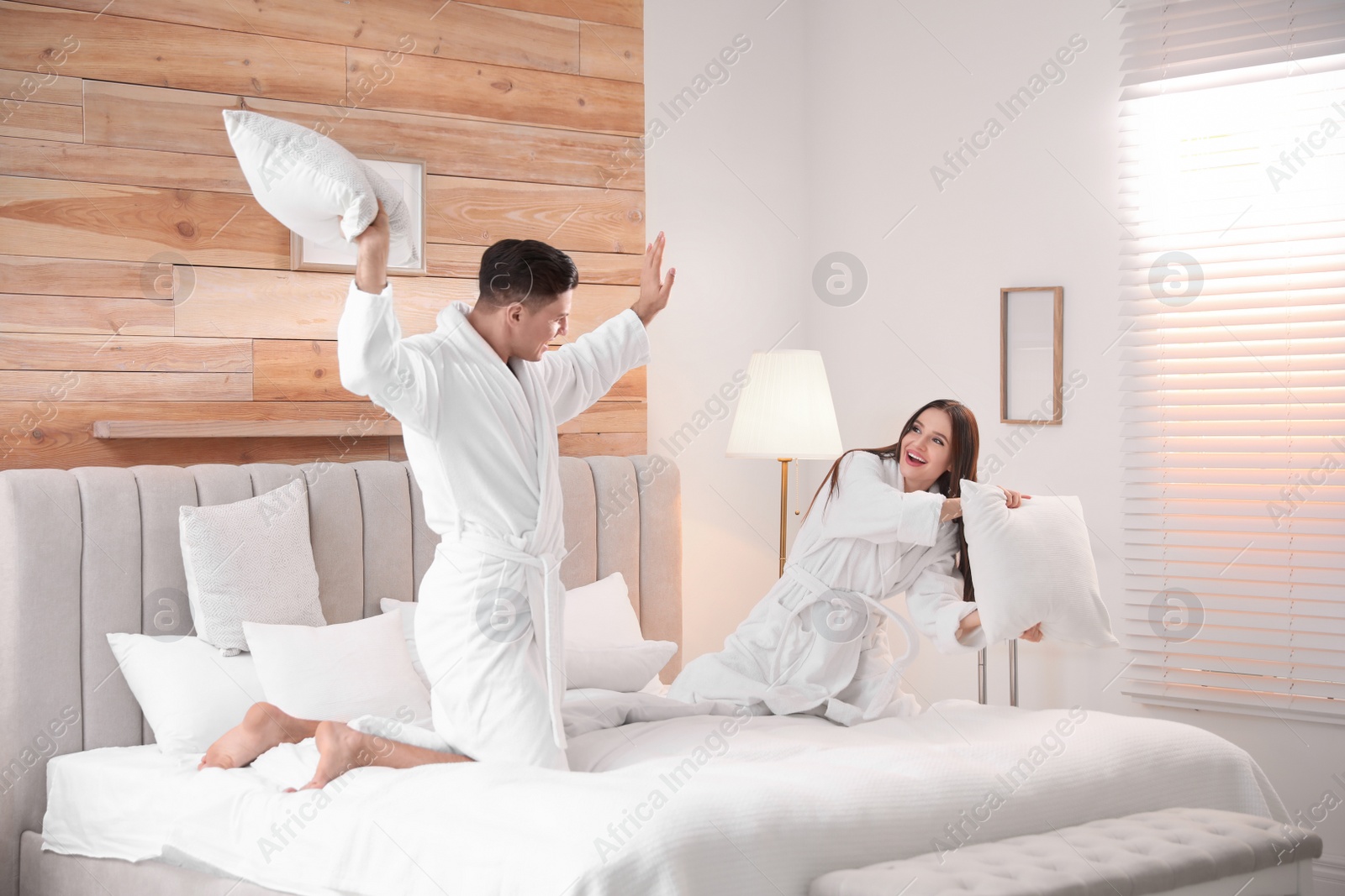 Photo of Happy couple in bathrobes having pillow fight on bed at home