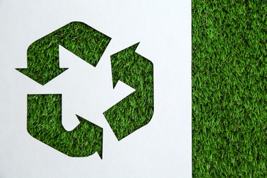 Photo of Sheet of paper with cutout recycling symbol on green grass, top view. Space for text