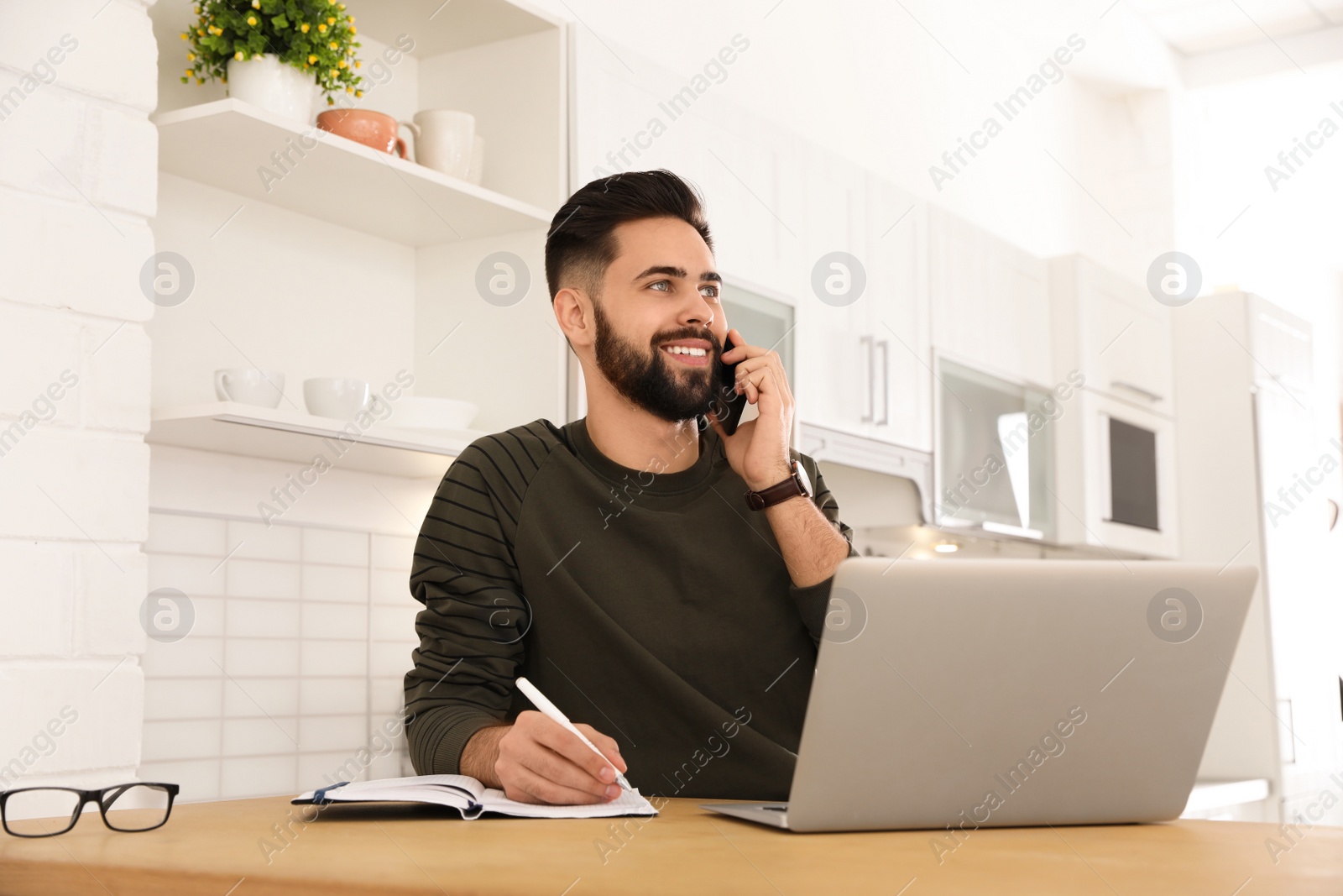 Photo of Handsome young man talking on phone while working at table in kitchen