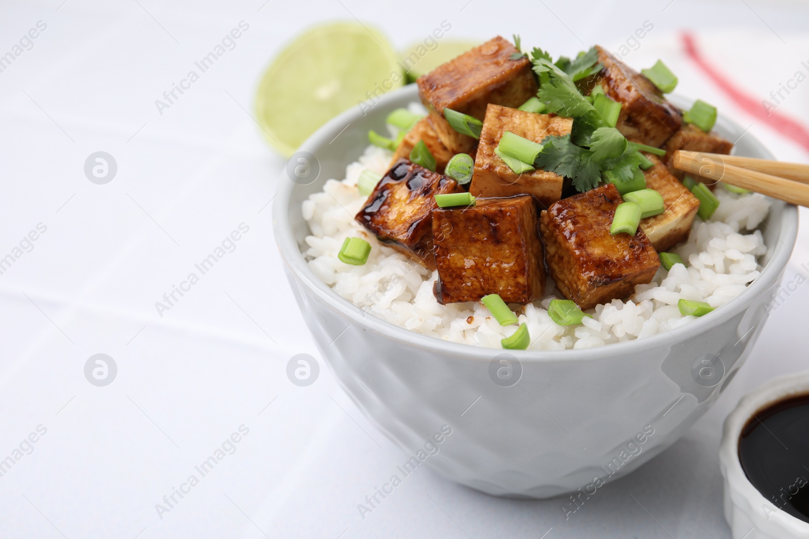 Photo of Bowl of rice with fried tofu and greens on white tiled table, closeup. Space for text