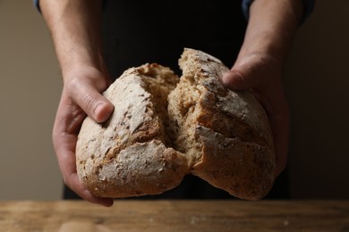 Photo of Man breaking loaf of fresh bread at wooden table, closeup