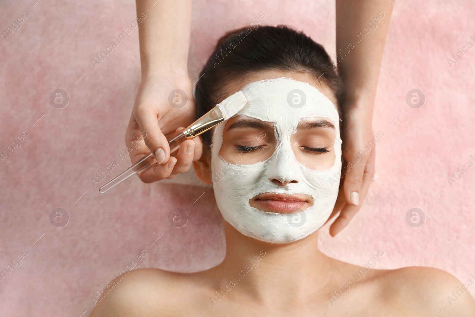 Photo of Cosmetologist applying white mask onto woman's face in spa salon, top view