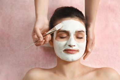 Cosmetologist applying white mask onto woman's face in spa salon, top view