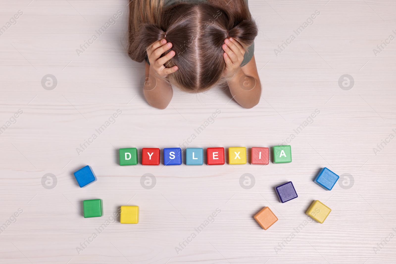 Photo of Little girl covering head with hands and cubes with word Dyslexia at white wooden table, top view