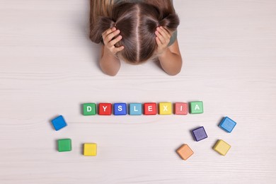 Little girl covering head with hands and cubes with word Dyslexia at white wooden table, top view