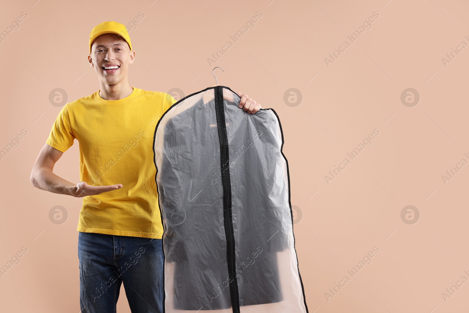 Photo of Dry-cleaning delivery. Happy courier holding garment cover with clothes on beige background, space for text