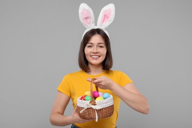 Photo of Easter celebration. Happy woman with bunny ears and wicker basket full of painted eggs on grey background