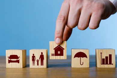 Man arranging cubes with different icons in row on wooden table against light blue background, closeup. Insurance concept