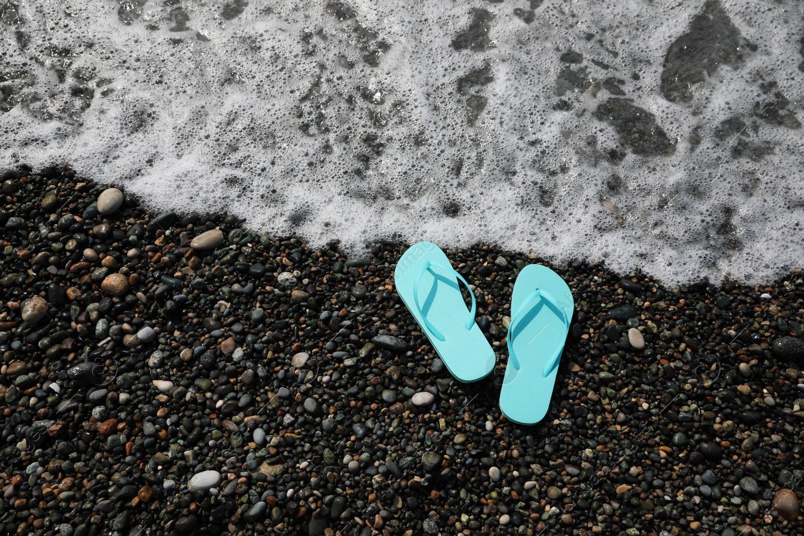 Photo of Stylish turquoise flip lops on pebble beach near beautiful sea wave, above view. Space for text