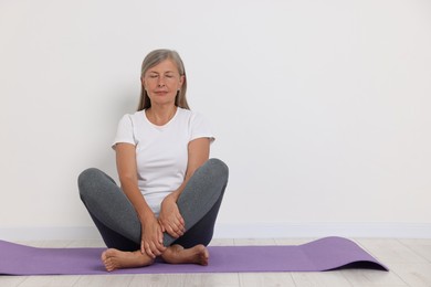 Senior woman practicing yoga on mat near white wall. Space for text