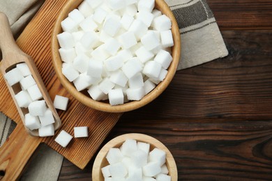 Photo of White sugar cubes in bowls and scoop on wooden table, flat lay. Space for text