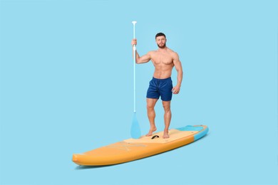 Handsome man with paddle on SUP board against light blue background