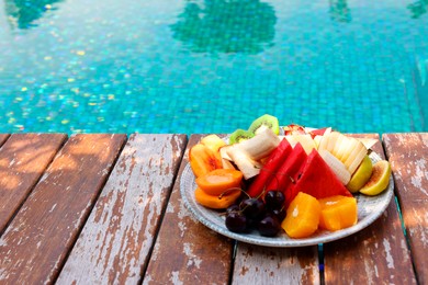 Photo of Plate with fresh fruits on wooden deck near outdoor swimming pool, space for text. Luxury resort