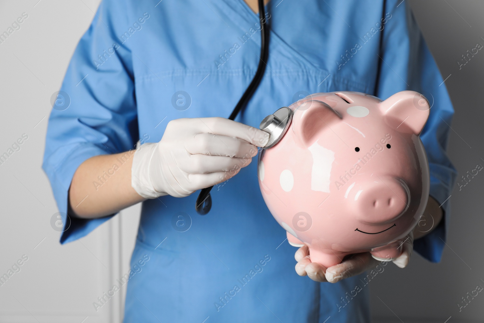 Photo of Doctor holding pale pink ceramic piggy bank with stethoscope against white wall, closeup. Medical insurance