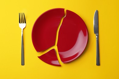 Pieces of broken red ceramic plate and cutlery on yellow background, flat lay