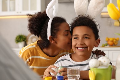 Photo of Happy African American mother kissing her cute son while painting Easter eggs at table in kitchen
