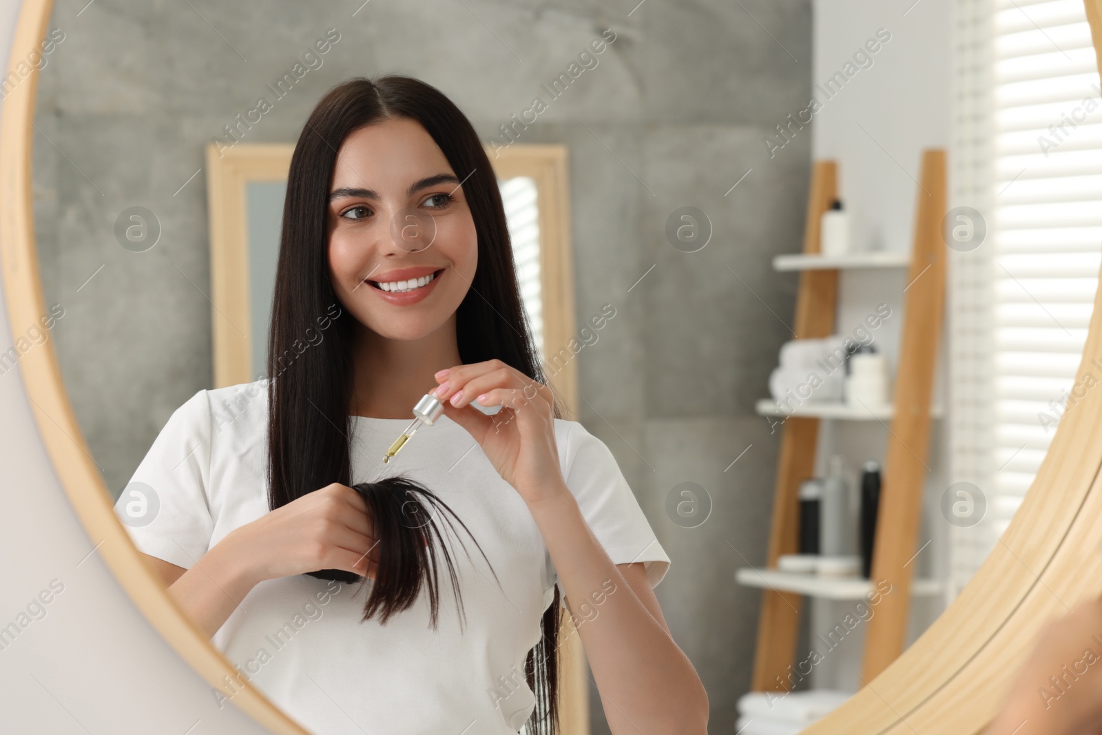 Photo of Beautiful woman applying hair serum in bathroom, reflection in mirror. Cosmetic product