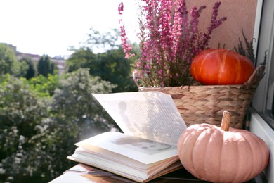 Photo of Wicker basket with beautiful heather flowers, pumpkins and open book on windowsill outdoors. Space for text