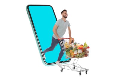 Image of Grocery shopping via internet. Happy man with shopping cart full of products running out of huge smartphone on white background