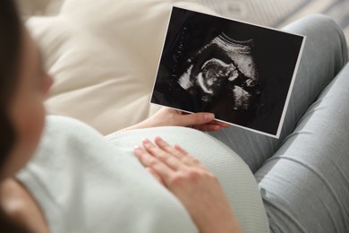 Pregnant woman holding ultrasound picture near her belly indoors, closeup