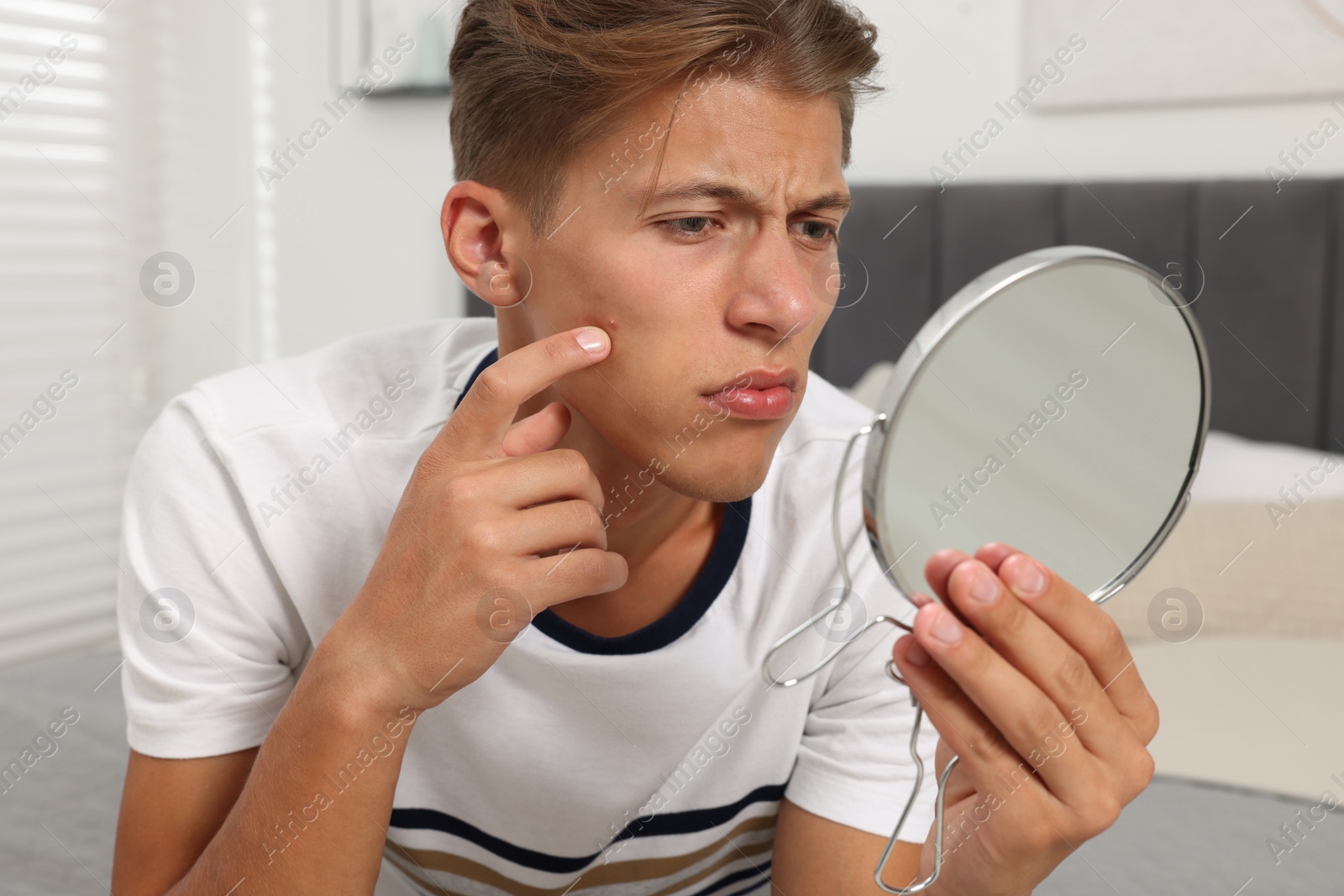 Photo of Upset young man looking at mirror and touching pimple on his face indoors. Acne problem