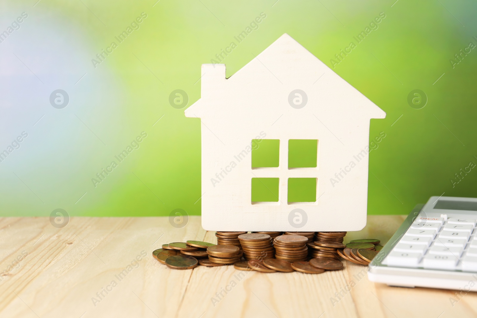 Photo of House model, calculator and coins on wooden table against blurred green background, space for text