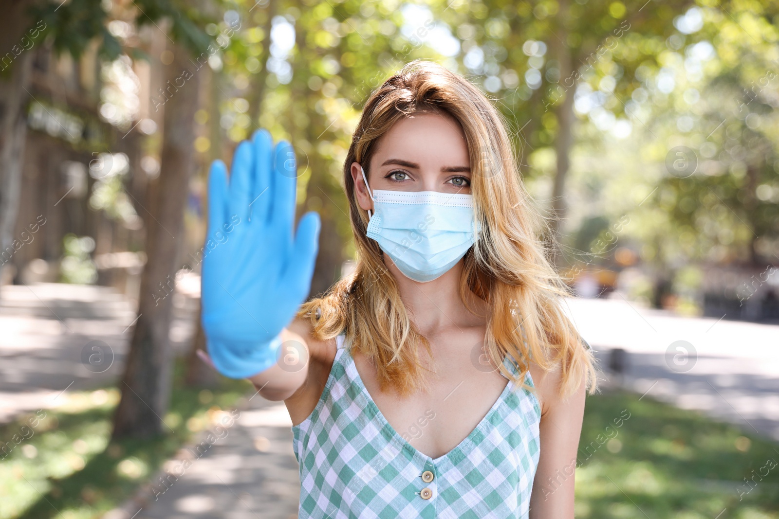 Photo of Woman in protective face mask showing stop gesture outdoors. Prevent spreading of coronavirus