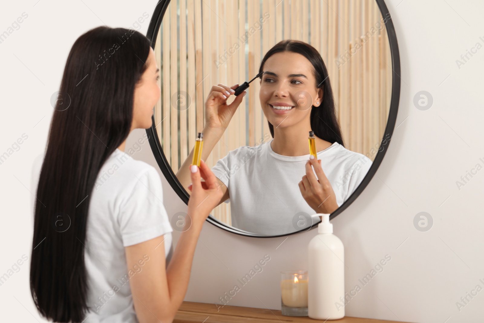 Photo of Young woman applying oil onto eyelashes near mirror indoors