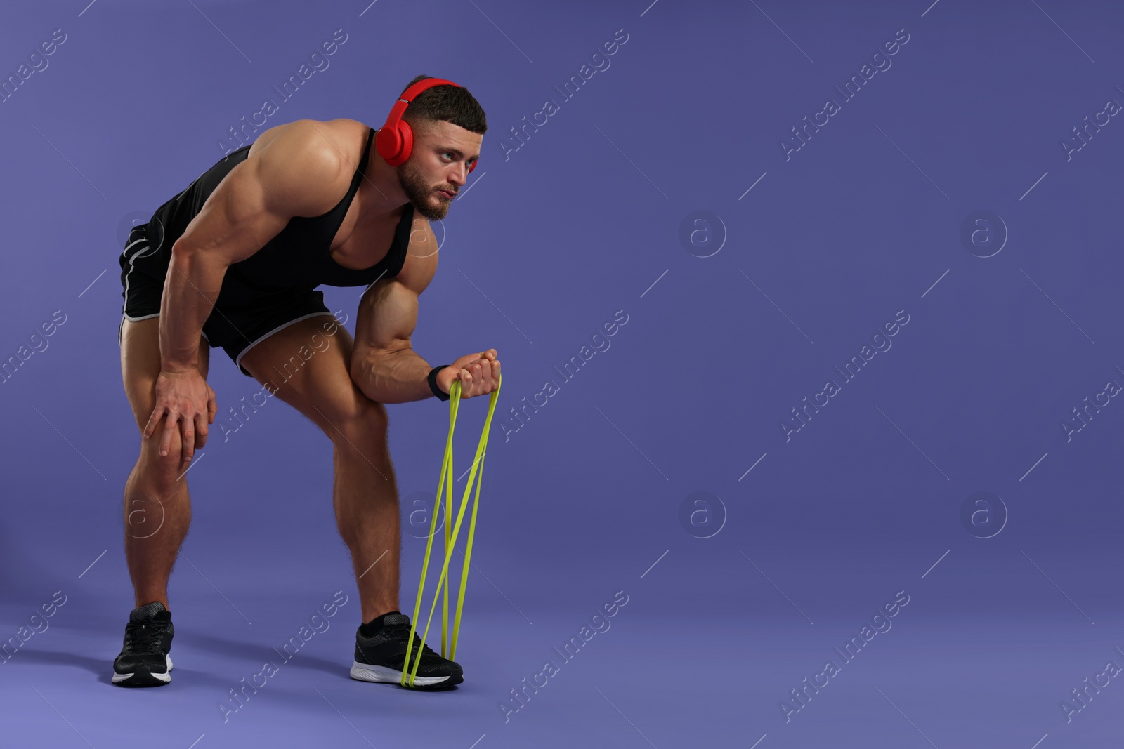 Photo of Muscular man in headphones exercising with elastic resistance band on purple background. Space for text