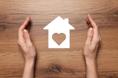 Image of Woman holding hands around paper silhouette of house on wooden table, top view. Insurance concept