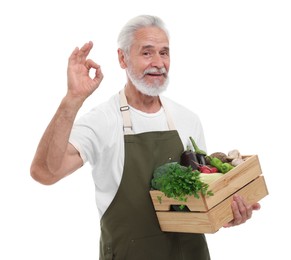 Harvesting season. Farmer holding wooden crate with vegetables and showing ok gesture on white background