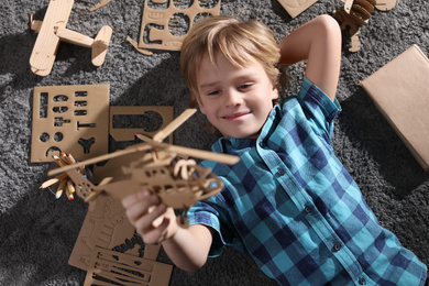 Photo of Little boy playing with cardboard helicopter on floor at home, top view. Creative hobby