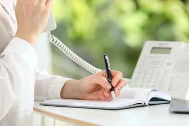 Photo of Assistant with telephone handset writing at white table against blurred green background, closeup