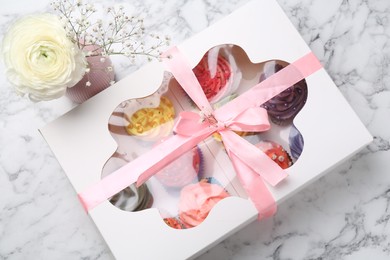 Different colorful cupcakes in box and flowers on white marble table, above view