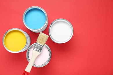 Photo of Cans of different paints and brush on red background, flat lay. Space for text