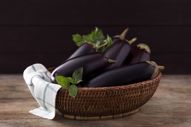 Photo of Ripe purple eggplants and basil in wicker bowl on wooden table, closeup
