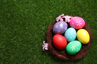 Colorful Easter eggs in decorative nest with flowers on green grass, top view. Space for text