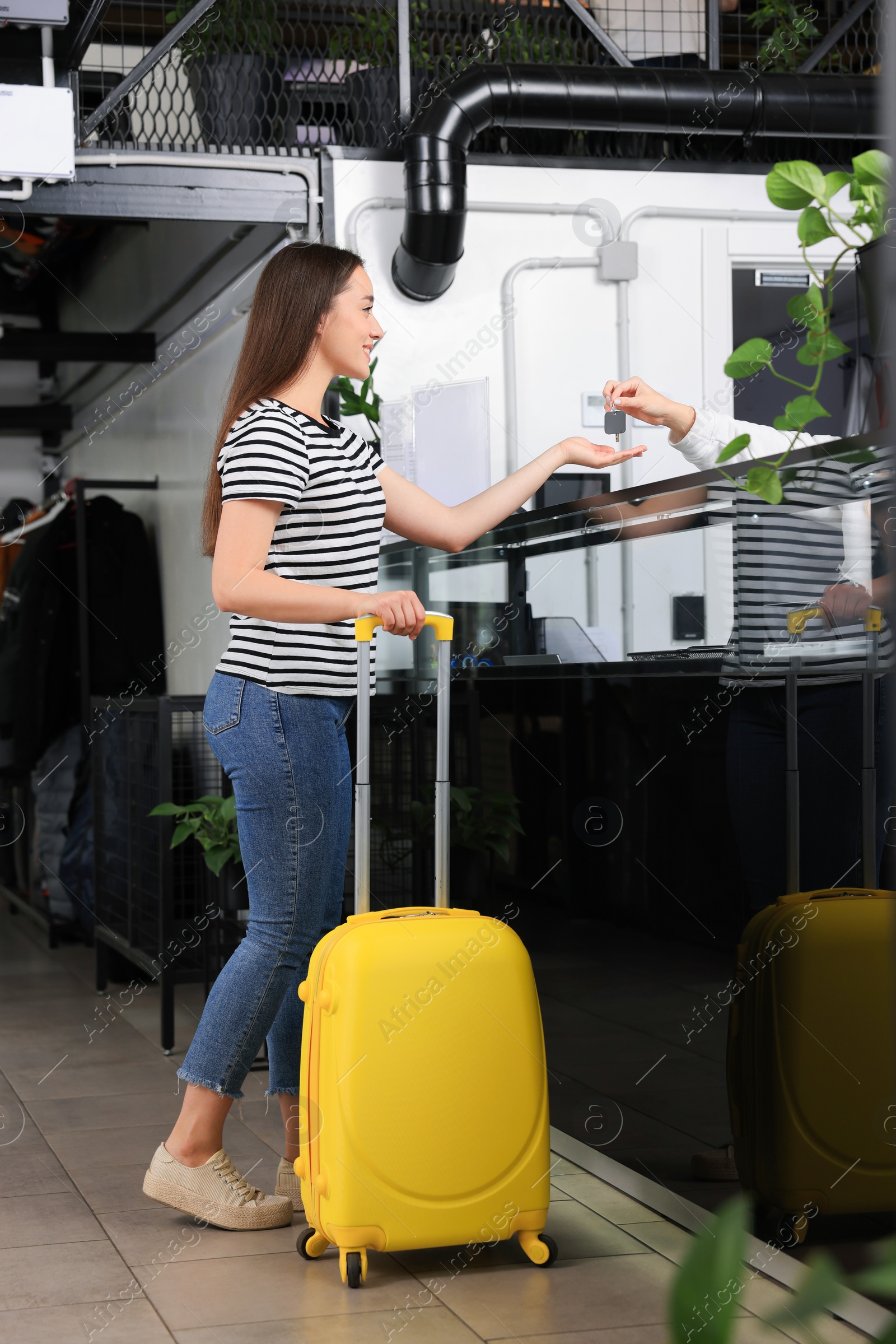 Photo of Happy young woman with suitcase receiving key at reception desk in hostel