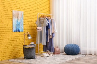 Rack with different stylish clothes and shoes near yellow brick wall in room