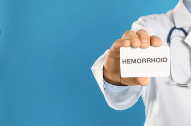 Photo of Doctor holding business card with word HEMORRHOID on blue background, closeup. Space for text