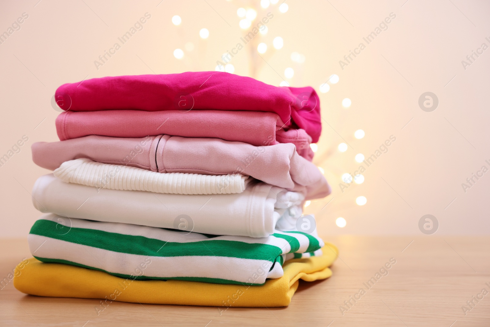 Stack of folded clothes on wooden table against beige background with blurred lights. Space for text Photo of Stack of folded clothes on wooden table against beige background with blurred lights. Space for text