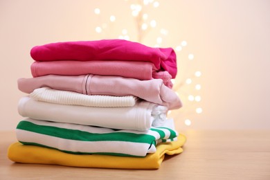 Photo of Stack of folded clothes on wooden table against beige background with blurred lights. Space for text