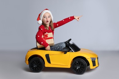 Photo of Cute little girl in Santa hat with Christmas gift box driving children's electric toy car on white background