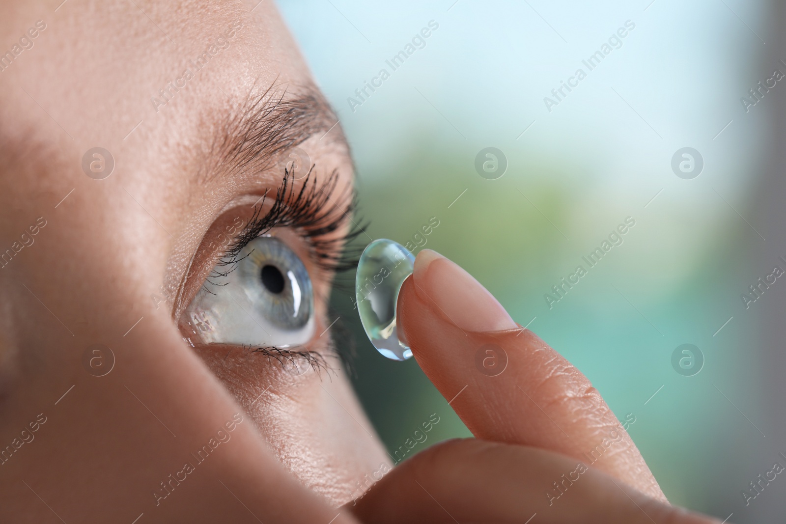 Photo of Young woman putting contact lens in her eye on blurred background, closeup