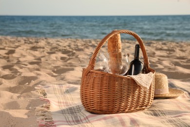 Blanket with bread, wine and glasses in basket for picnic near straw hat on sandy beach