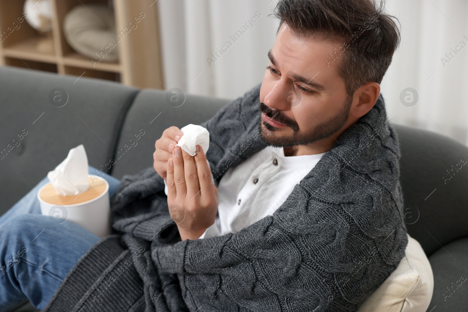 Photo of Sick man wrapped in blanket with tissue on sofa at home. Cold symptoms