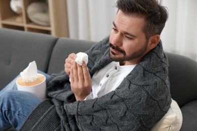 Sick man wrapped in blanket with tissue on sofa at home. Cold symptoms