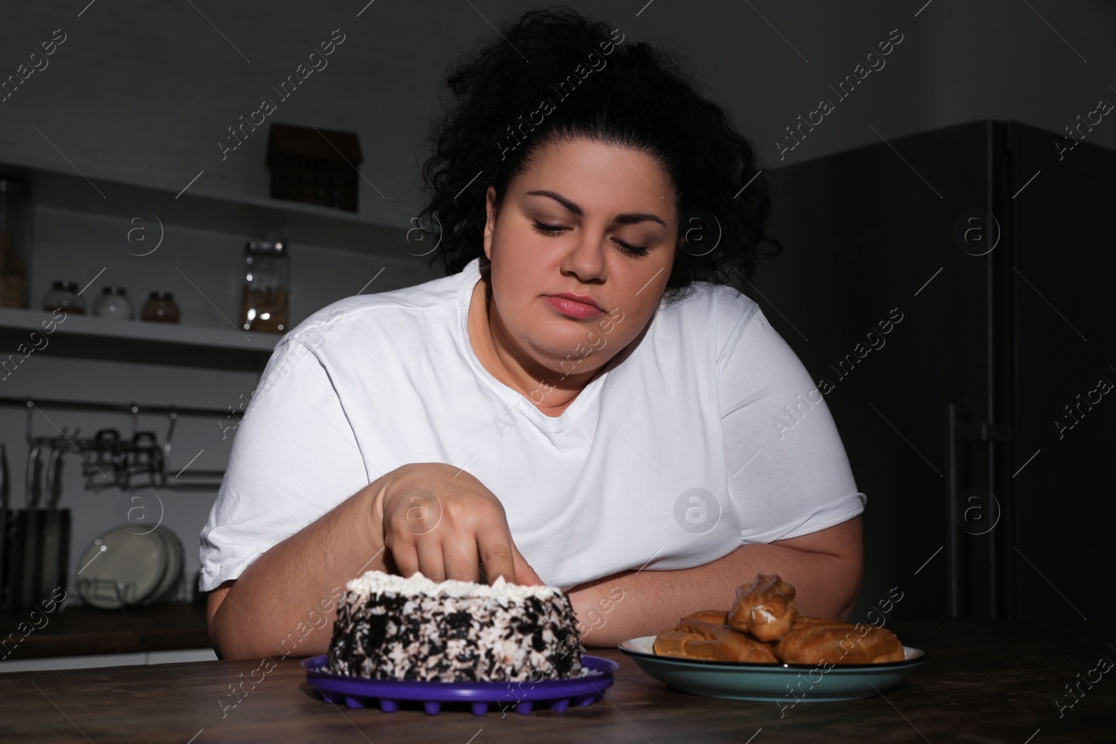 Photo of Depressed overweight woman eating cake in kitchen at night