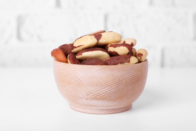 Bowl with tasty Brazil nuts on white table against brick wall, closeup